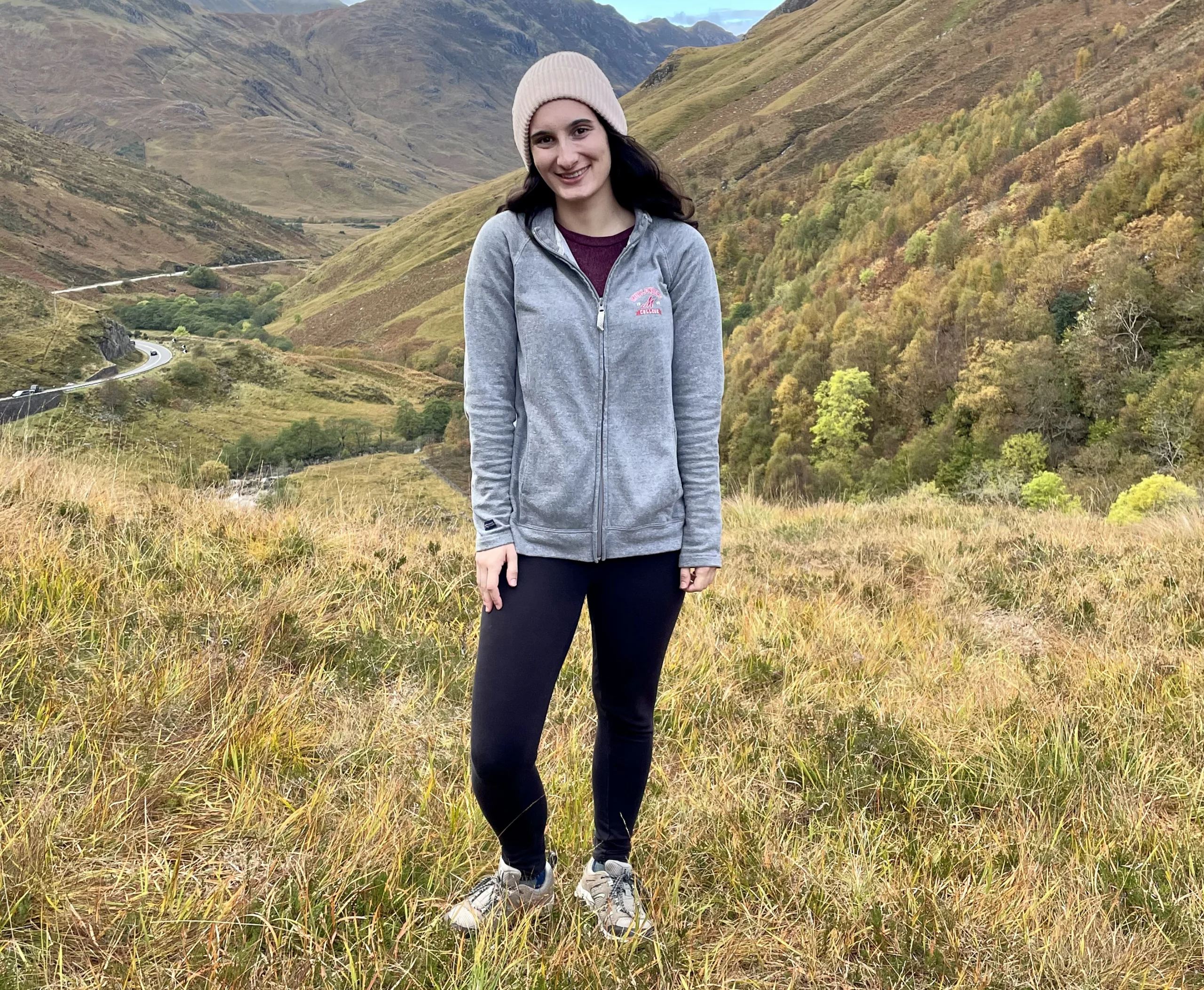 A young person stands in front of grassy mountains
