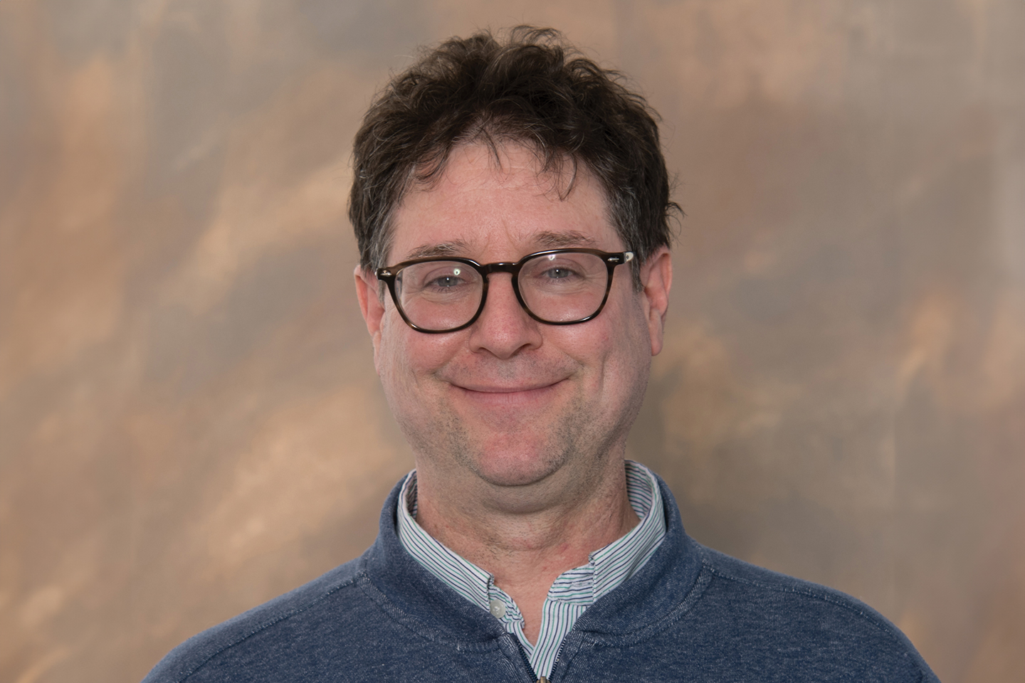 A headshot of a college professor in glasses on a brown background