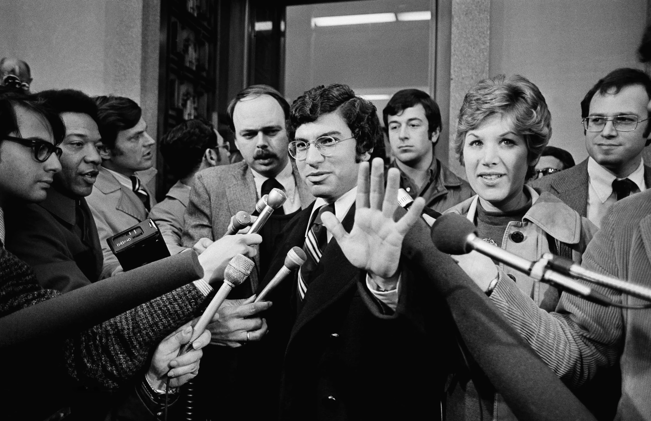 An archival black and white photo of a man in a suit holding up his hand while journalists hold microphones in his face