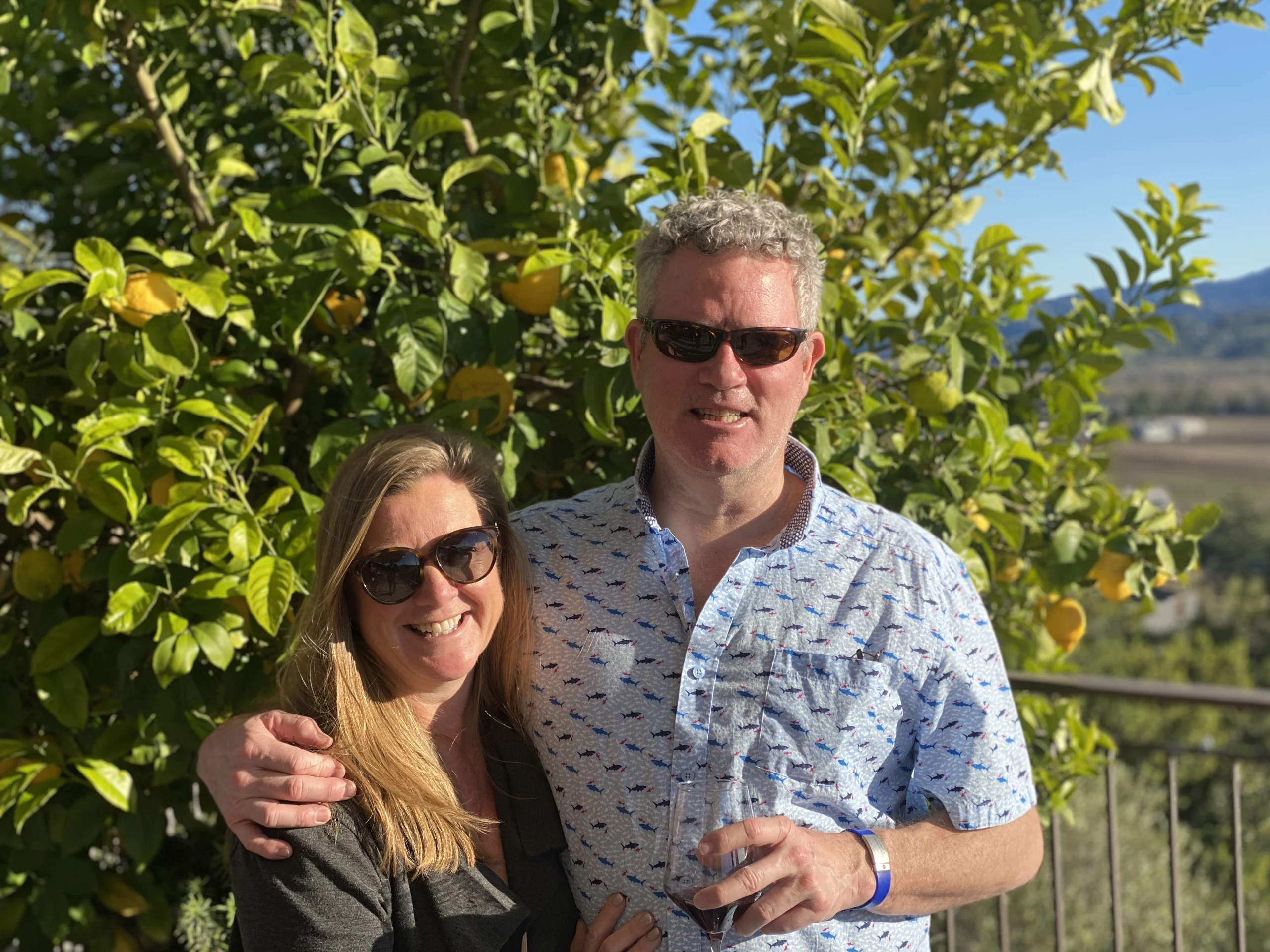 Two people stand in front of an orange tree while wearing sunglasses