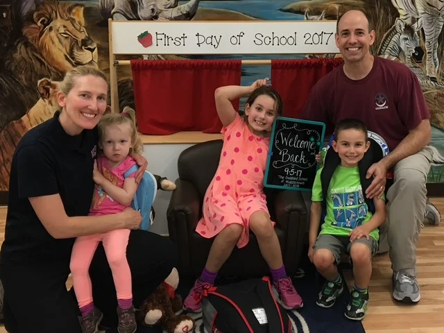 Two parents pose with three small children in front of a sign that says First Day of School 2017