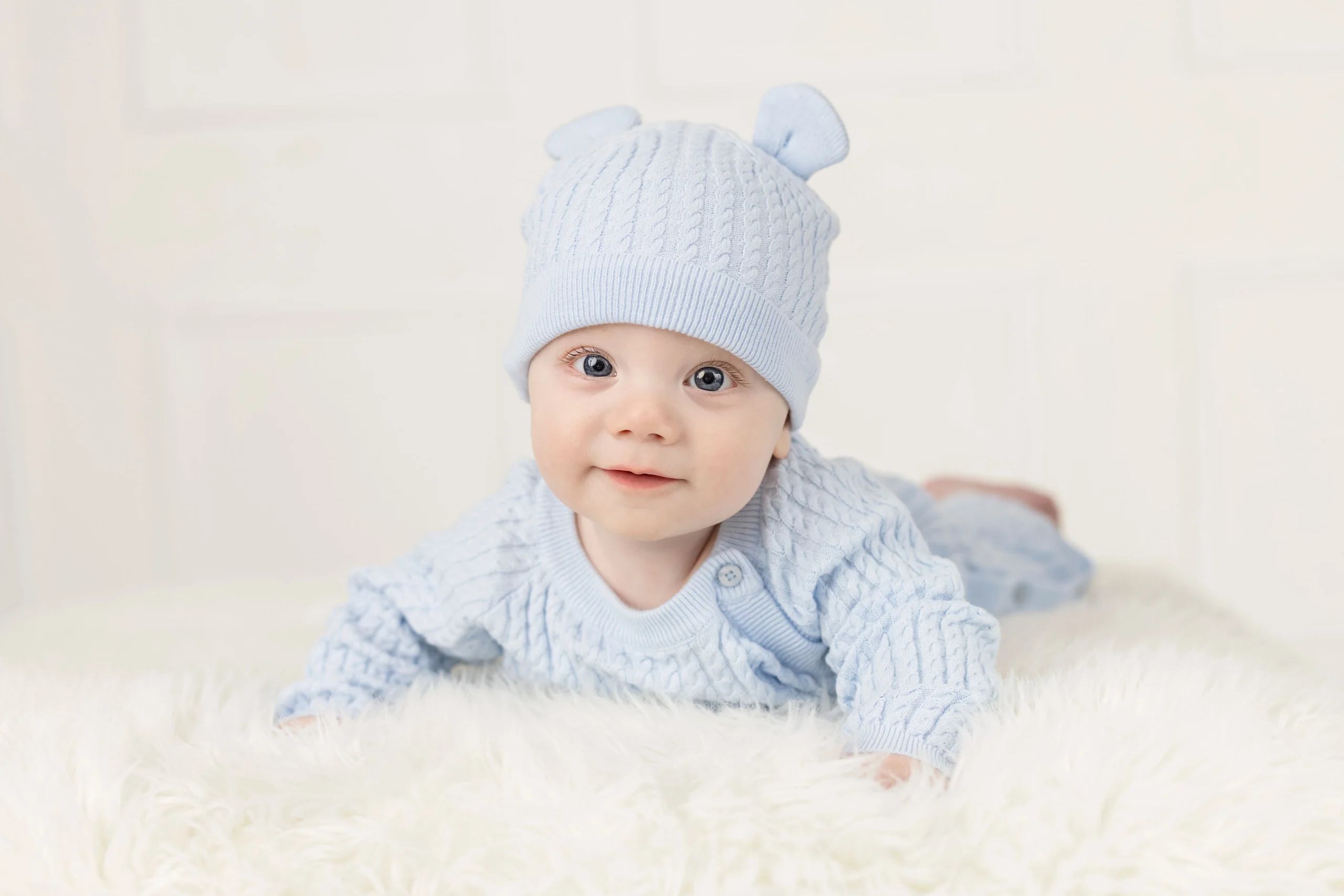 A baby in a blue outfit and hat lays on their belly for a portrait