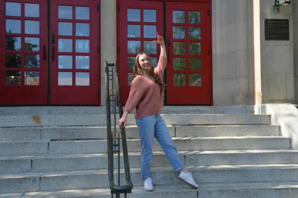 A college student wearing a pink sweater and jeans does a dance pose outside red doors