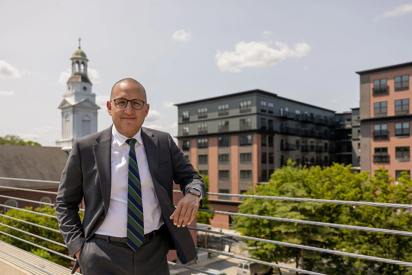 A man wearing glasses and a suit stands with city buildings in the background