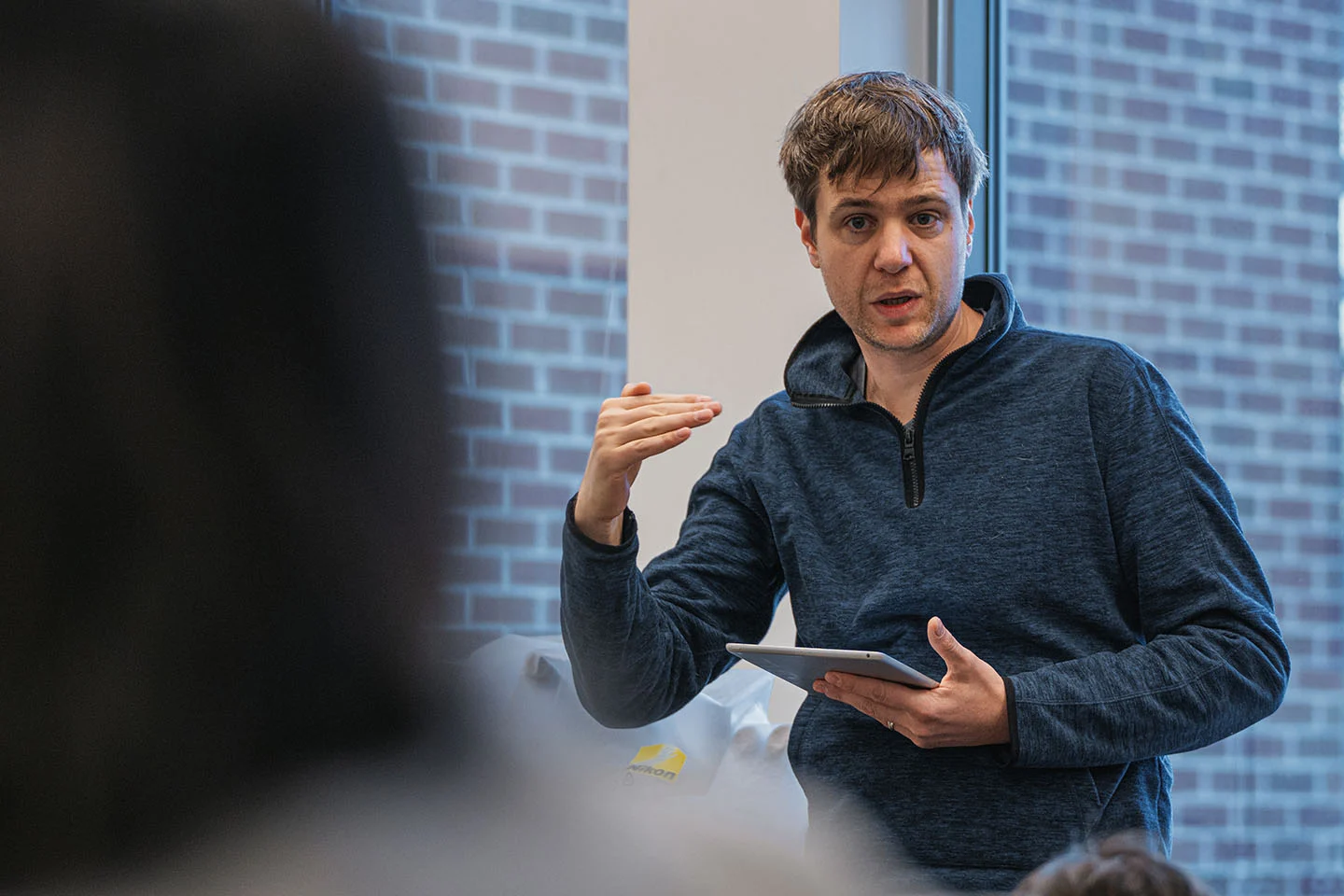A college professor in a blue shirt gestures as he speaks to students