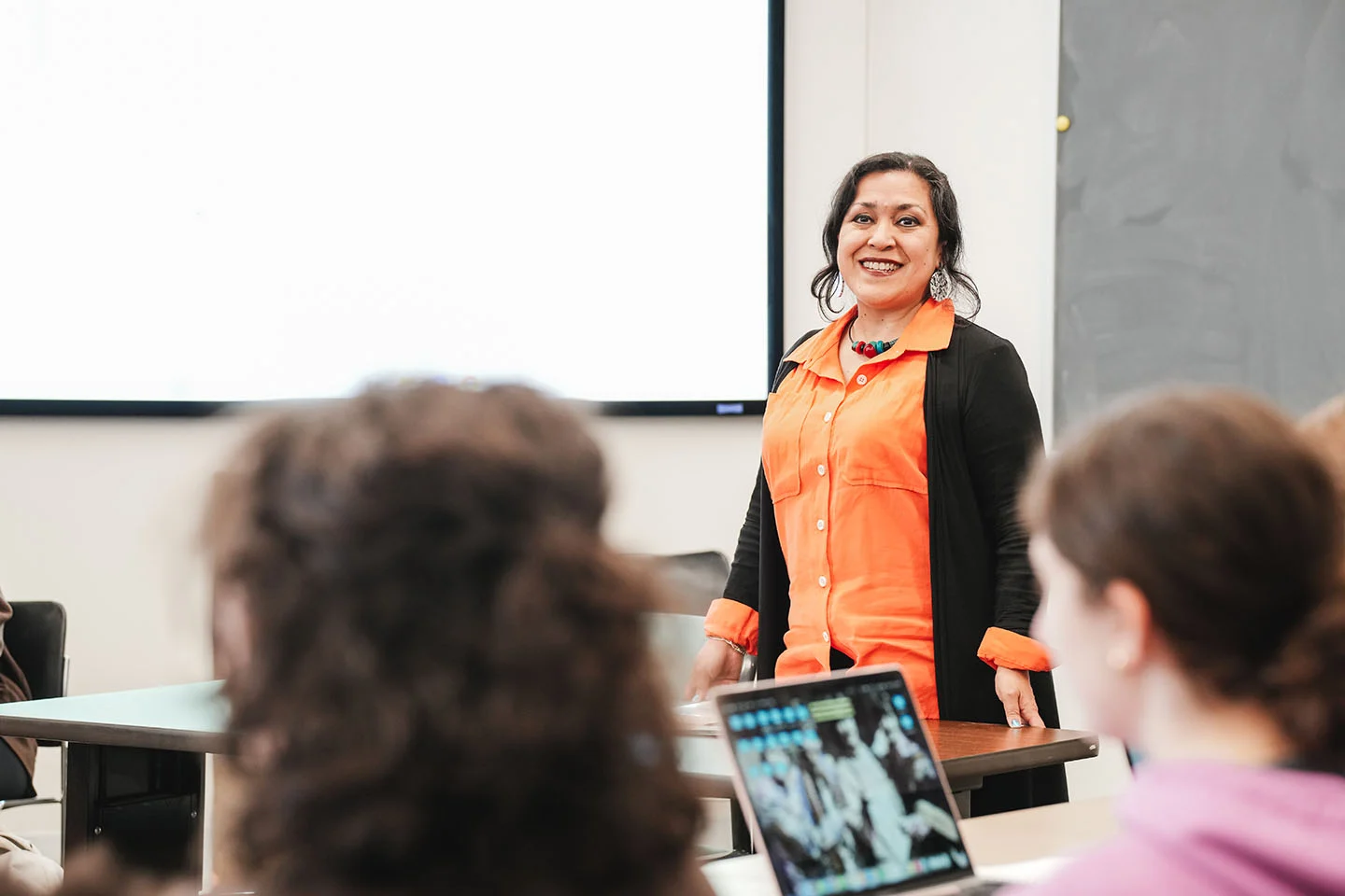A college professor in an orange shirt and black sweater smiles at her class